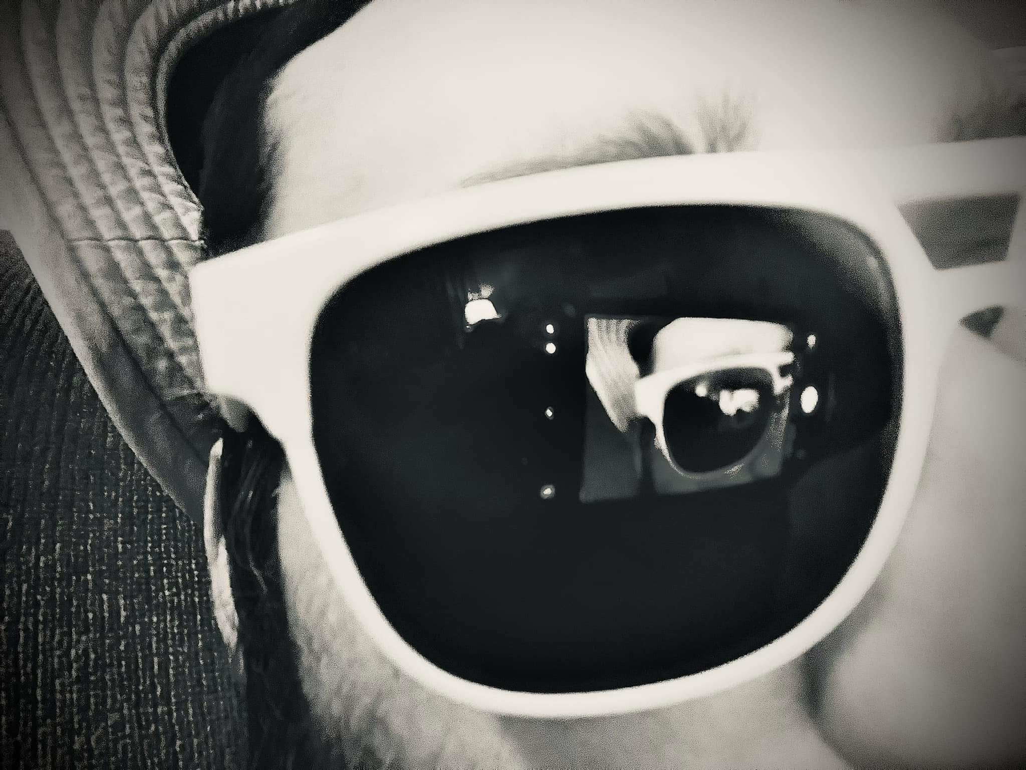 Black and white close-up of white sunglasses with a recursive reflection of the wearer in the lens.